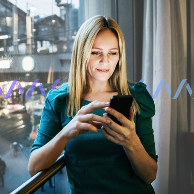 Photo of a woman standing by a window in a hotel room scrolling on her cell phone.