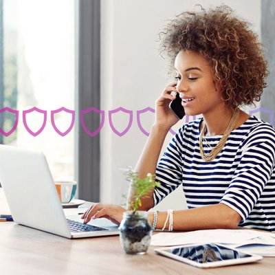 Photo of a woman in an office setting talking on her phone while typing or scrolling on her laptop. In the background, a series of small purple shields runs across the frame, representing security.