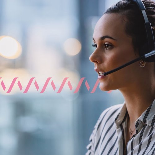 Photo of a contact center agent speaking to a customer through her headset. The woman is in the foreground and the background is blurred. A series of small pink wave forms run across the frame, representing communication.
