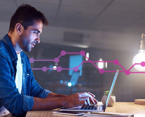 Photo of a man sitting at a table in an office setting typing on his laptop. Laying on the table in the foreground are a cell phone and a tablet. In the background, a series of purple lines and dots runs across the frame.