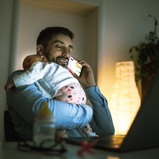 Photo of a business man at home, talking on his cellphone. He is holding a sleeping infant over his shoulder.