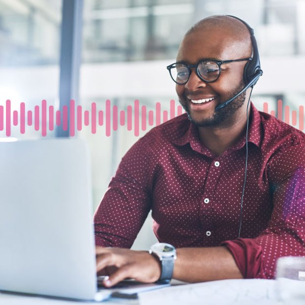 Photo of a smiling agent in an office typing on his laptop while speaking to a customer on his phone headset. In the background, a series of small pink vertical lines runs across the frame.
