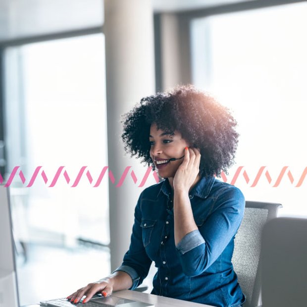 Photo of a smiling woman in an office talking into her headset and looking at her computer. In the background, a series of small wave-like icos run across the frame, representing communication.