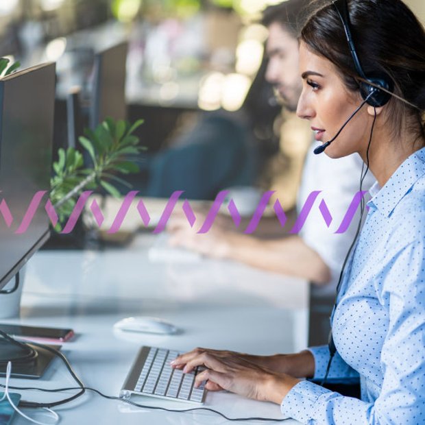 Photo of agents working in a call center. In the foreground, a woman is typing on her computer while talking to a customer on her headset. In the background, a series of small purple waves runs across the frame.