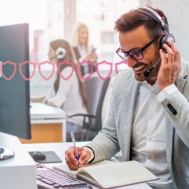 Photo of a smiling agent writing in a notebook while talking to a customer on his headset. In the background, a series of small pink shield icons runs across the frame., representing security