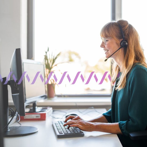 Photo of a smiling woman in an office typing on her desktop computer while talking to someone on her phone headset. In the background, a series of small purple waves run across the frame, representing communication.