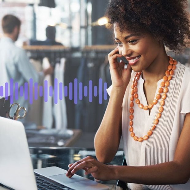 Photo of a smiling employee in a store. She is talking on the phone while scrolling on her computer. In the background, a series of small purple vertical lines run across the frame, representing communication.