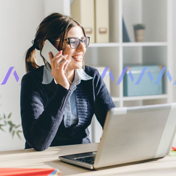 Photo of smiling woman sitting in front of her computer and talking to a customer. In the background, a small purple wave pattern runs across the frame, representing communication.