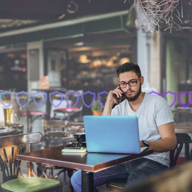Photo of a casually dressed man sitting alone at a table in a restaurant working on his laptop and talking on his cell phone. Running across the frame are a series of small shield icons representing security.