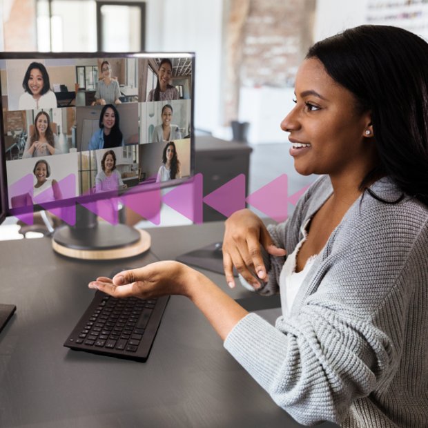 Photoillustration of a woman sitting in front of a desktop computer with a video conference reflected on her computer screen; the woman is using her left hand to gesture toward the screen, indicating she is part of the conference